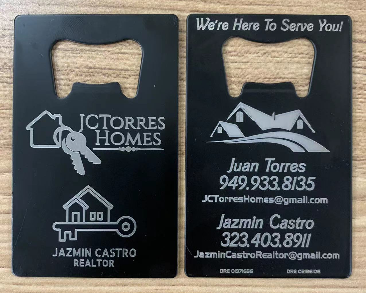 Two black metal bottle openers with JCTorres Homes branding on a wooden surface.