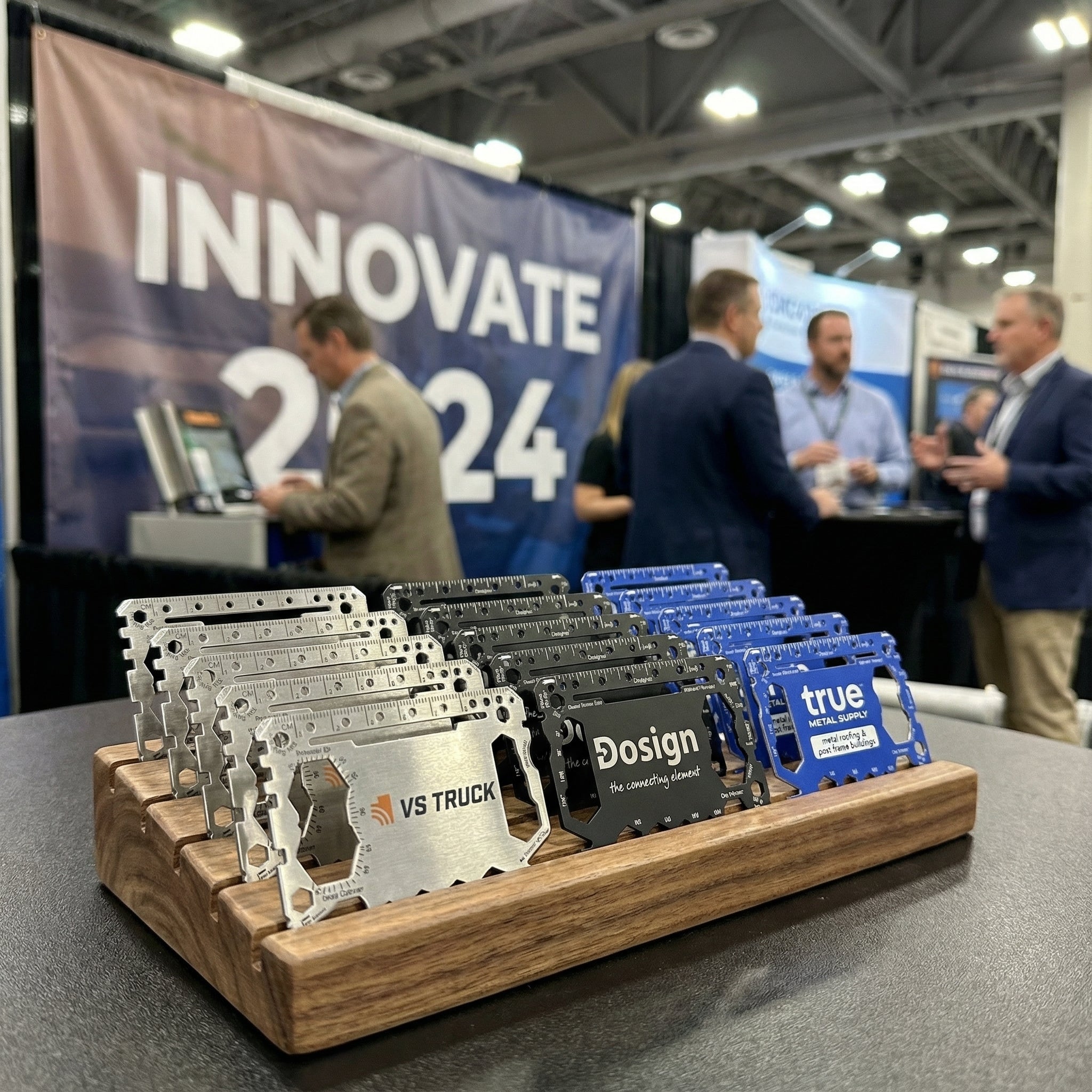 Display of multiple custom branded multi tools on a wooden stand with people and a 'Innovate 2024' banner in the background.